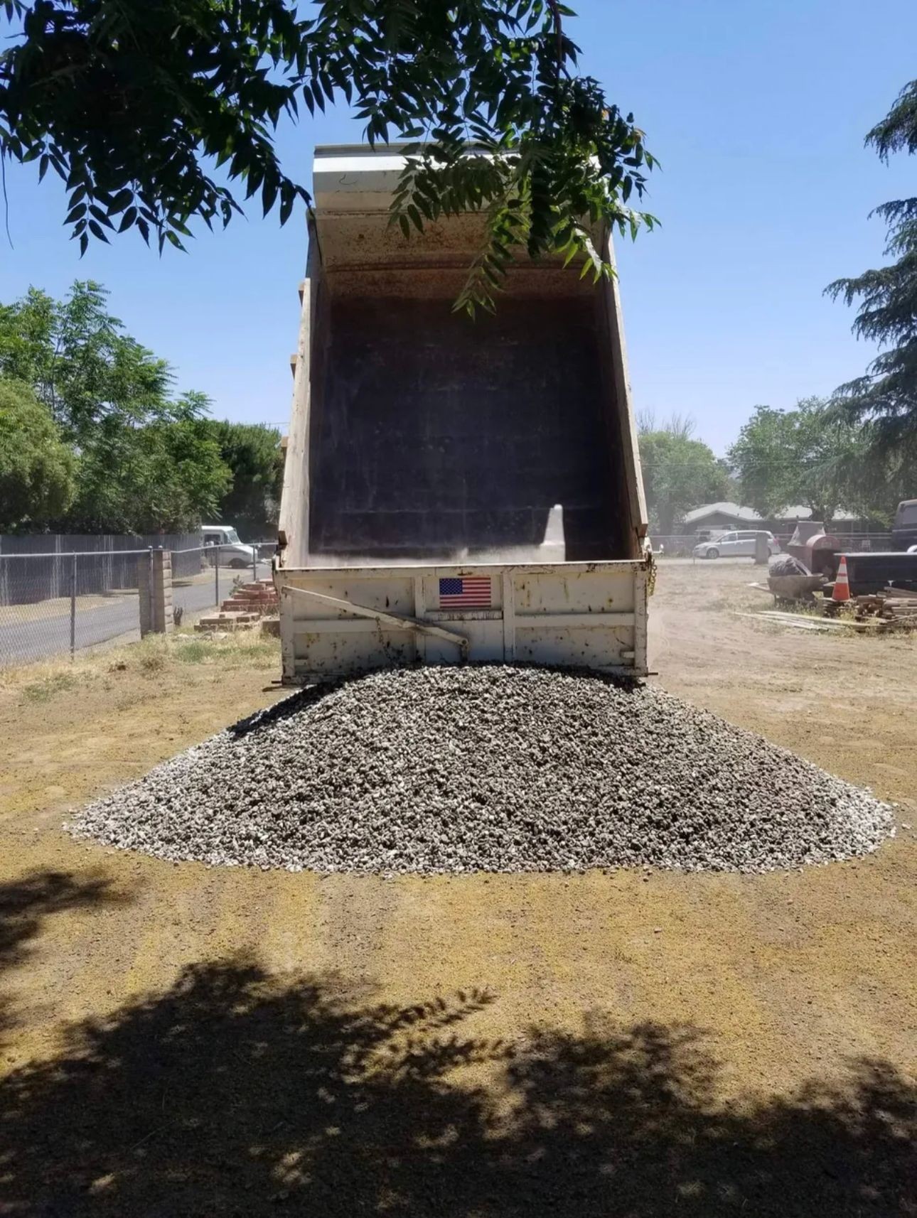 Dump truck unloading a pile of gravel on a dirt surface, surrounded by trees and blue sky.