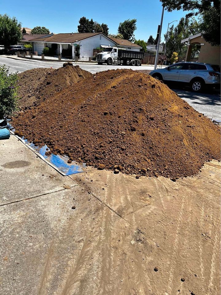 Large pile of dirt on a residential street with a dump truck and houses in the background.