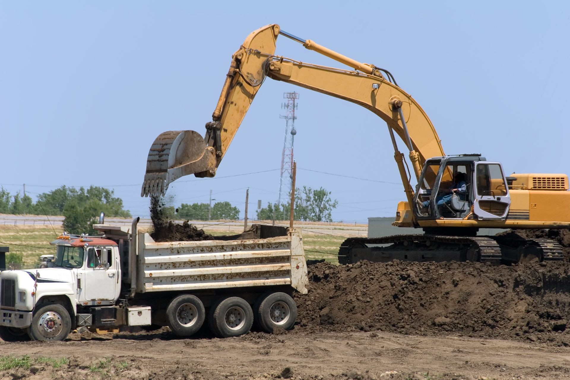 Excavator loading soil into a dump truck on a construction site under a clear blue sky.