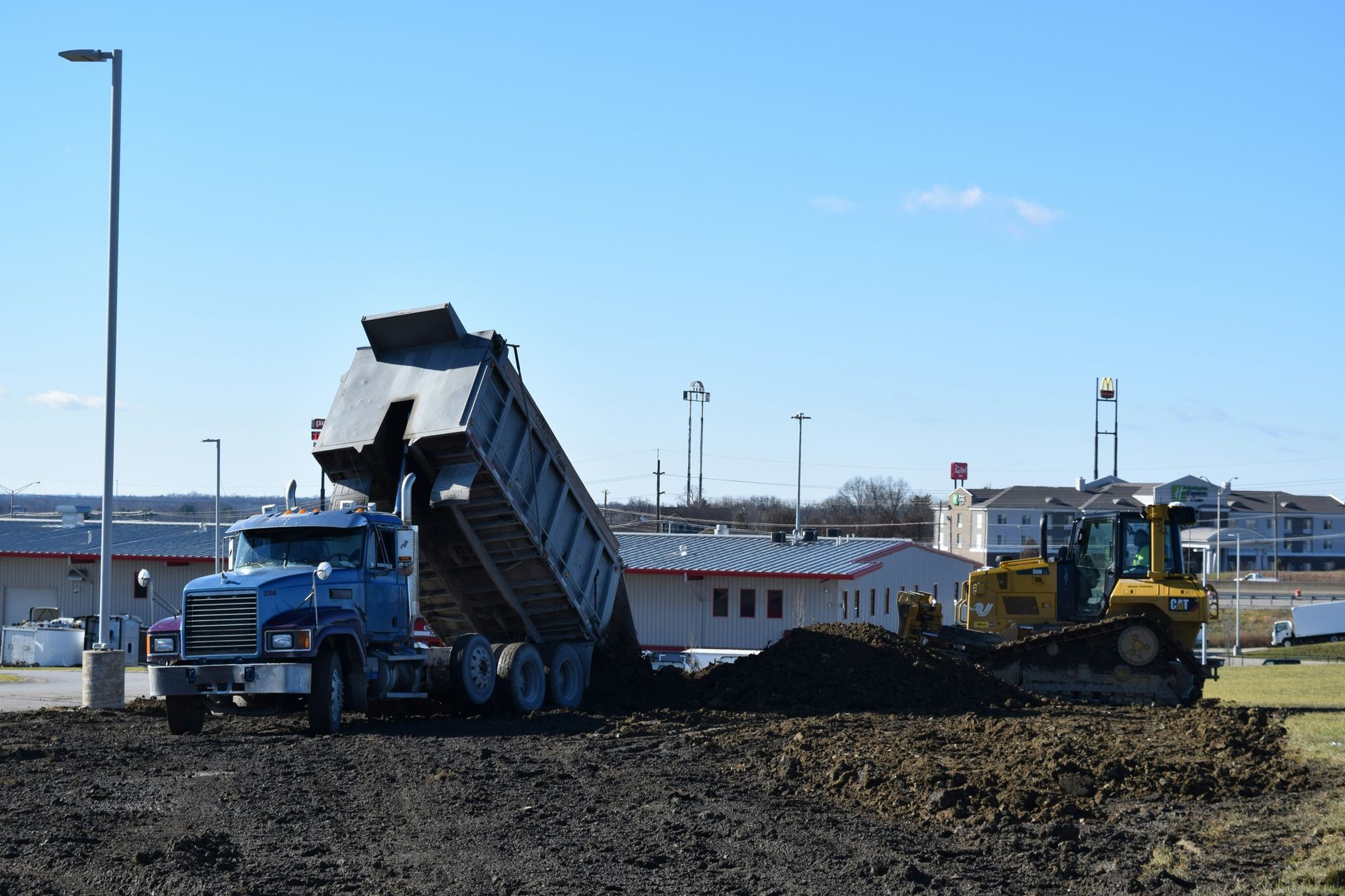 Dump truck unloading soil near a bulldozer at a construction site on a clear day.