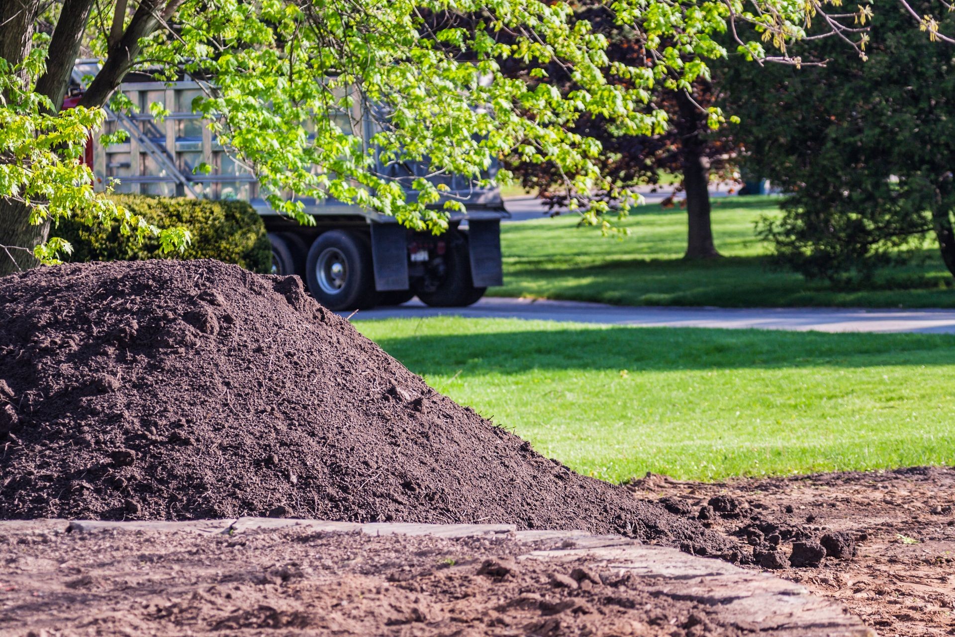 Pile of fresh soil in a park, with greenery and a truck in the background.