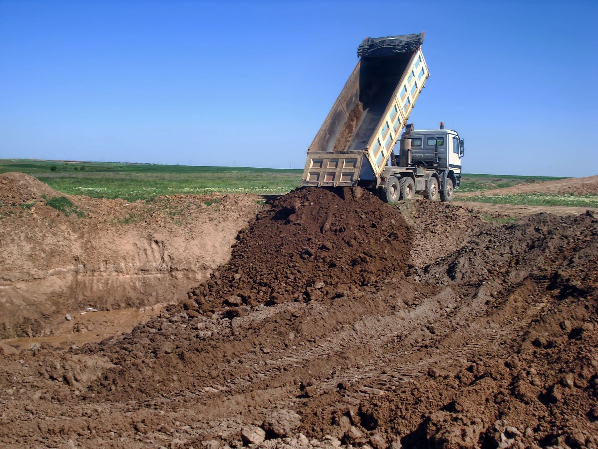 Dump truck unloading dirt at a construction site in a rural area under a clear blue sky.